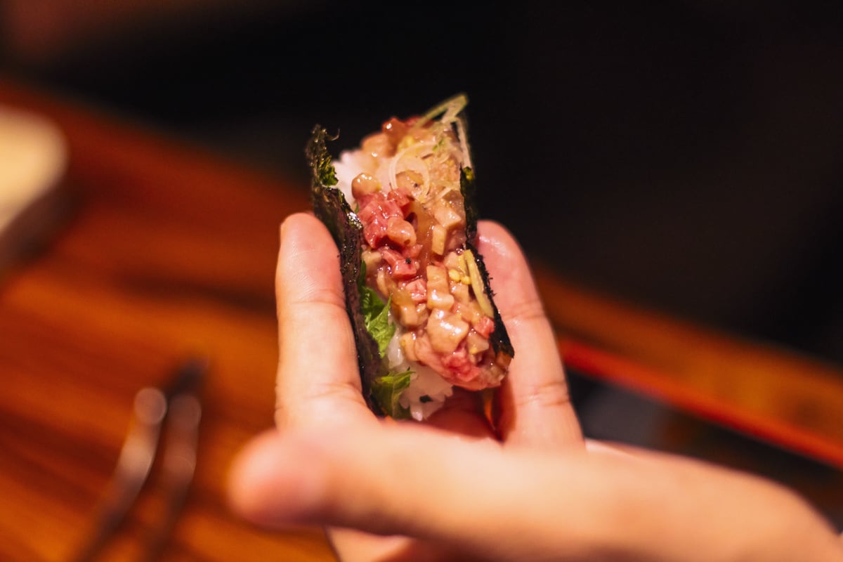 「黒タンのトロたく」を左手で持つ写真。海苔の上にご飯・大葉・黒たん・ネギが乗っている