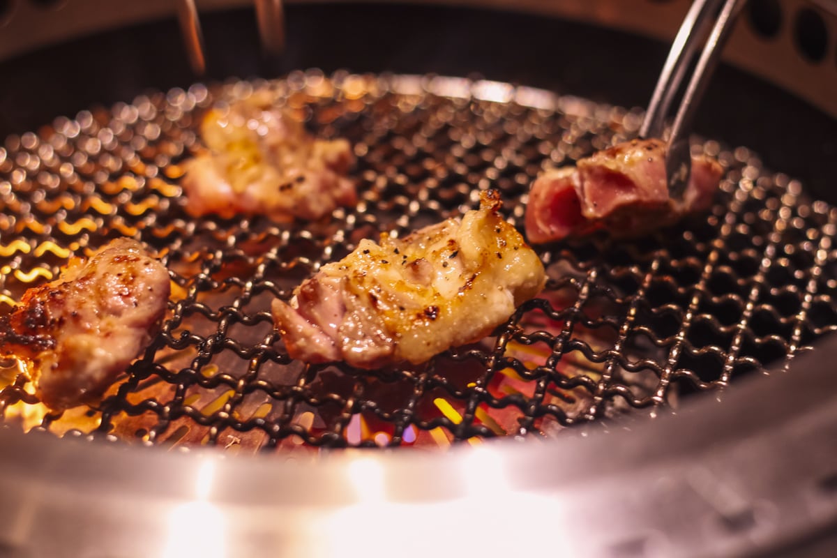 網の上に4切れの名古屋コーチンが並んでいて、右側の肉をトングで掴んでいる様子