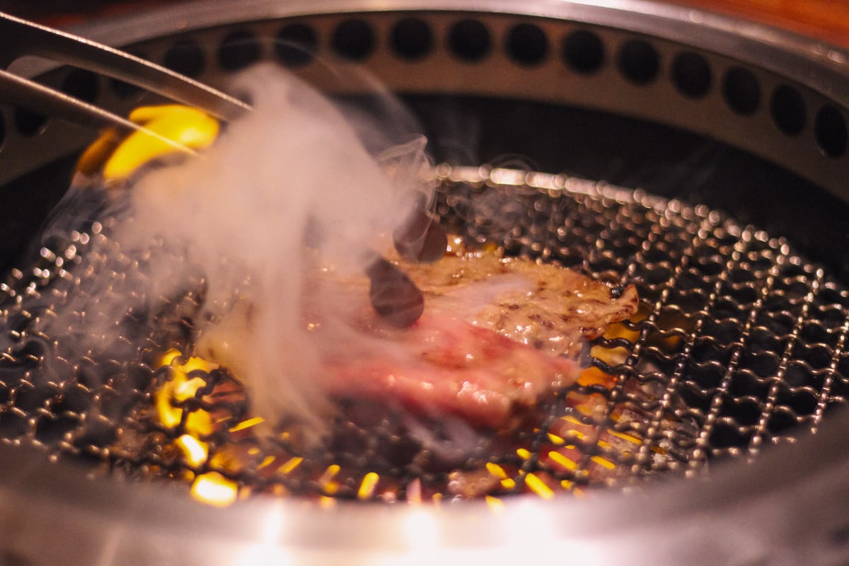 網の上でサーロインのスライスを広げ、トングでひっくり返す様子。左側に白い煙があがっている