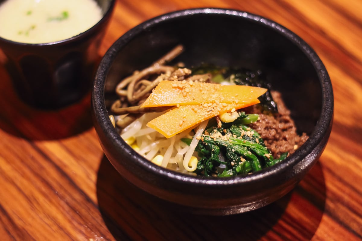 黒く分厚い小さめのどんぶりに盛られたビビンバ、左奥に黒い器に玉子スープが注がれている