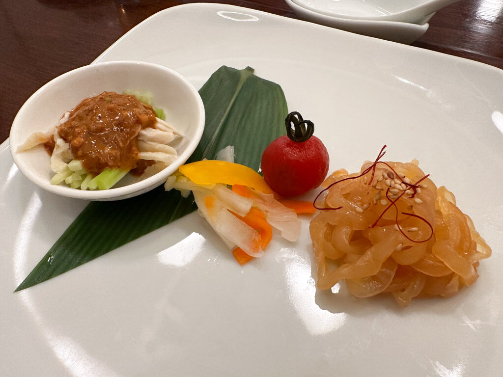 左に白いお皿にバンバンジーが盛り付けられている。中央にはピクルスとプチトマト、右側にクラゲが盛り付けられている。